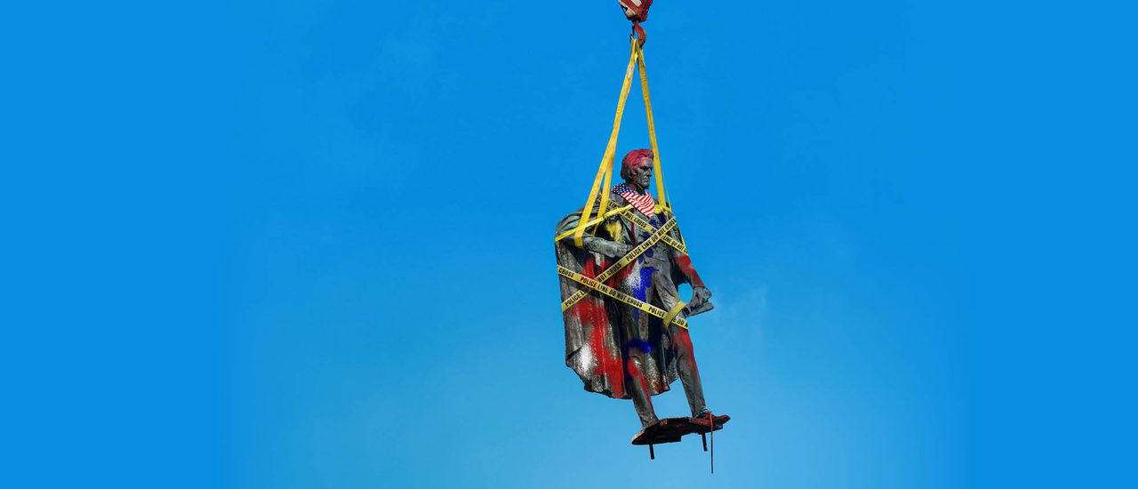 A traditional stone statue of a historic figure is suspended from an orange metal hook with yellow straps against a bright blue background. It is wrapped in yellow 'police line do not cross' tape and defaced with red, white and blue graffiti and an American flag neck scarf.