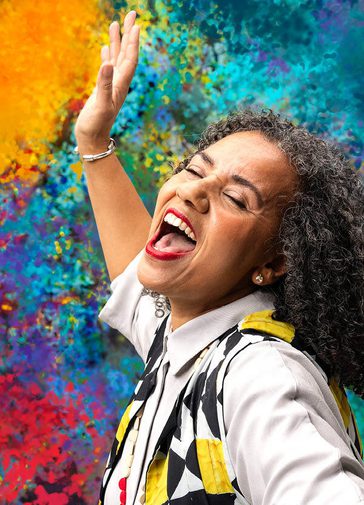 A woman sings joyfully, eyes closed and mouth open wide. Her head is passionately thrown back and her arm is raised. She has black and grey curly hair and wears a white short sleeved shirt with a black, yellow and white patterned waistcoat and bright red lipstick. The background is a bright array of colourful, dappled paint.