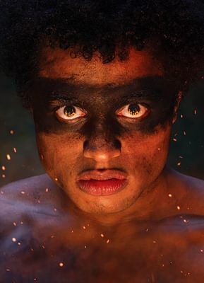 A young man is looking directly into the camera, with a serious look on his face. Flames are reflected in his eyes and sparks surround him.