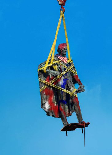 A traditional stone statue of a historic figure is suspended from an orange metal hook with yellow straps against a bright blue background. It is wrapped in yellow 'police line do not cross' tape and defaced with red, white and blue graffiti and an American flag neck scarf.
