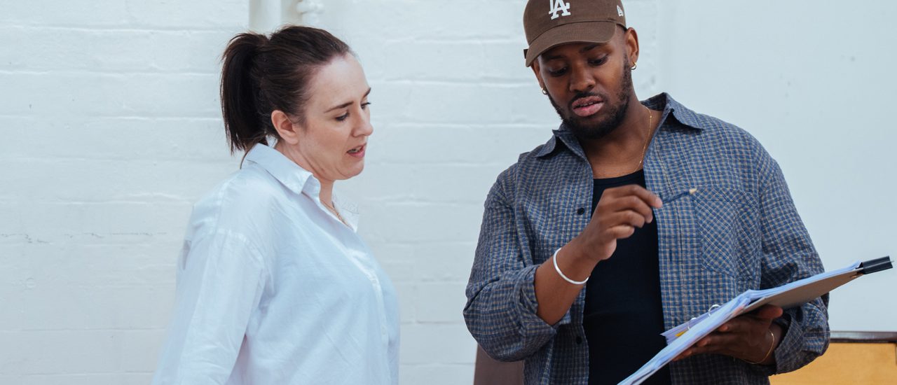 A man and woman in a rehearsal room. They are both reading from a document he is holding.