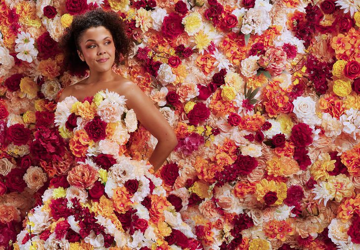 A young woman stands totally surrounded by flowers. They flood the background and cover her strapless, floor-length gown. The flowers are a mixture of pink, white, yellow and orange tones. The woman holds her hand behind her back and looks off to the side with a content, quietly confident facial expression.