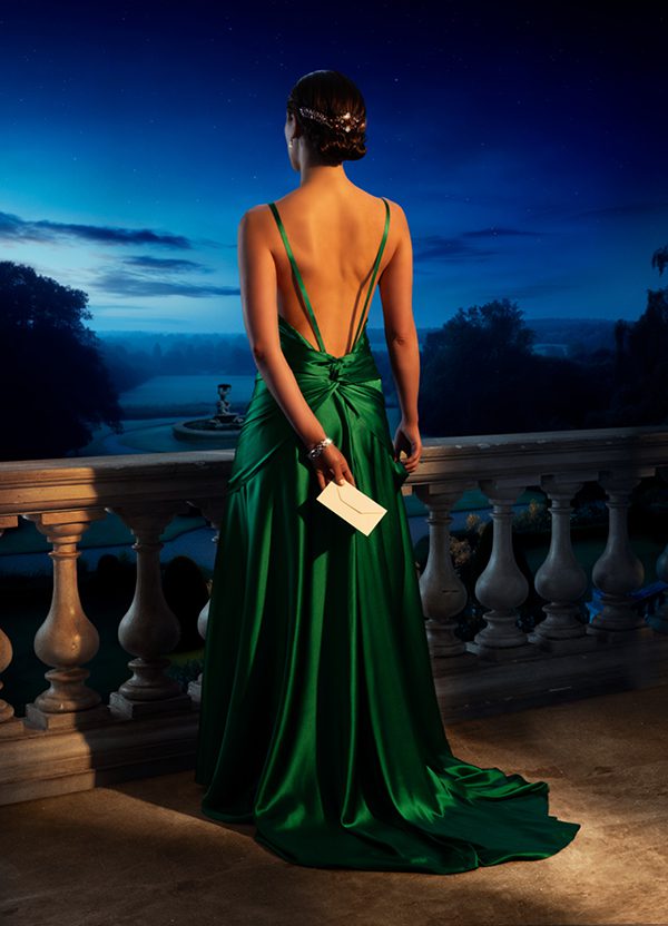 A young woman in a striking green backless dress holds a white envelope in one hand behind her back. She looks out at a garden and fountain from a balcony at dusk.