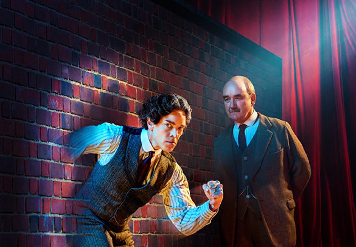 A dark-haired man with a determined expression is running through a brick wall. A stark spotlight is shining directly on him. A man with a neat moustache is looking at him intently with his hands behind his back.