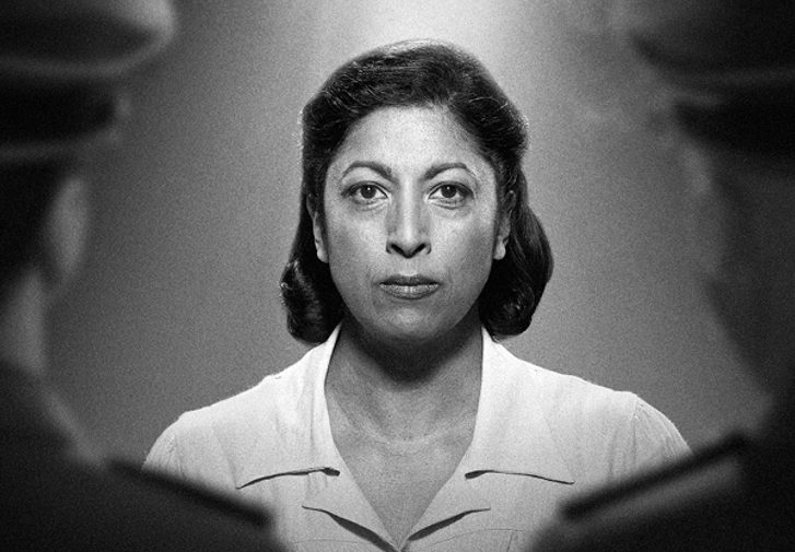 In a black and white photograph, a woman with dark shoulder-length hair stares resolutely at the camera, wearing a white top. A light illuminates her from above. Slightly out of focus, facing her on either side, are two men in military uniform with their backs to the camera.