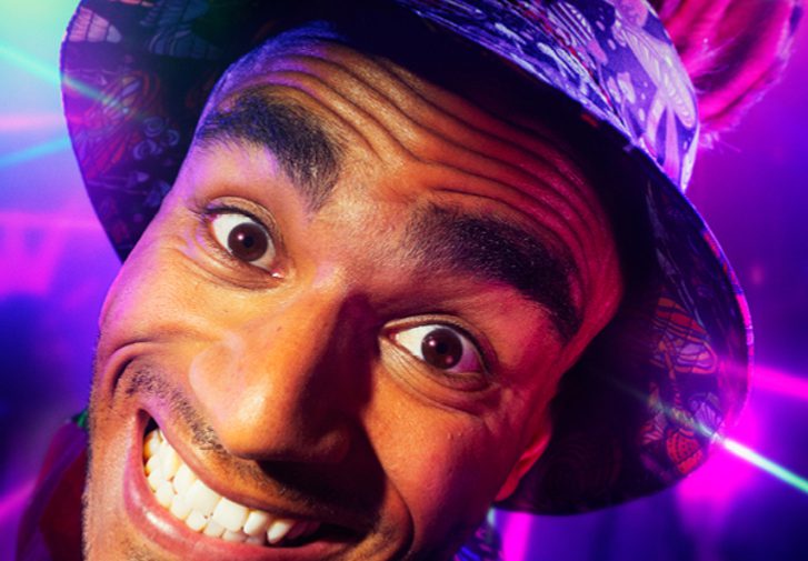 A young man is grinning intensely at the camera. He is wearing a bucket hat with donkey ears. The background is a colourful rave with purple and green strobe lights.