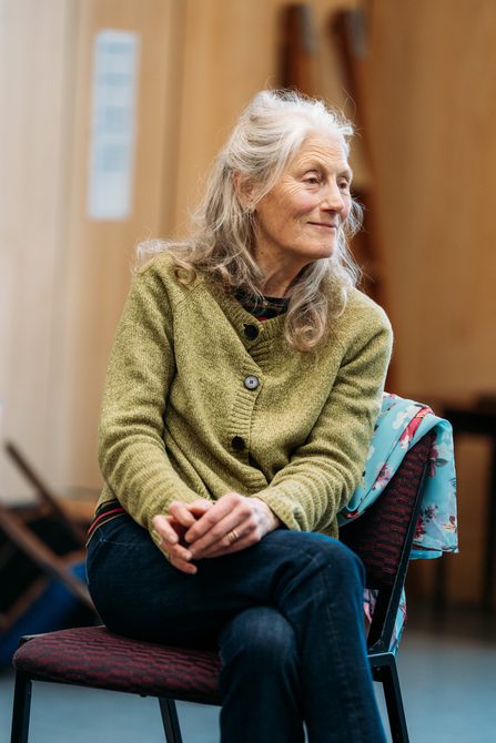 An actress leans forward on a chair, she smiles wistfully.