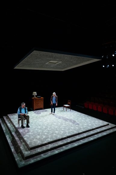 A man and woman are on a minimal set. The floor is a large carpeted square, above them is a smaller square suspended from the ceiling. She stands, facing towards him, as he sits in a chair with a book in his hands.