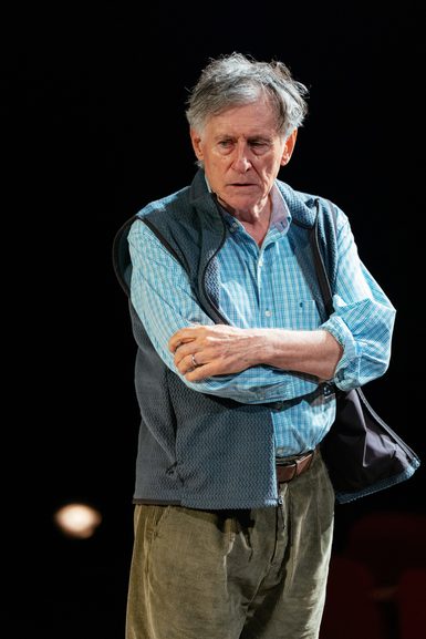 An older man is stood with his arms folded and a dismayed expression. He wears corduroy trousers, a blue check shirt and a gilet.