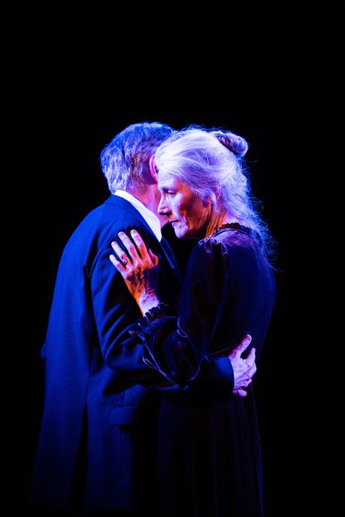 An older man and woman are stood in an embrace. She looks out with a contemplative expression, while his face is covered by hers. They are both wearing formal black clothing. A blue light from above creates atmosphere.