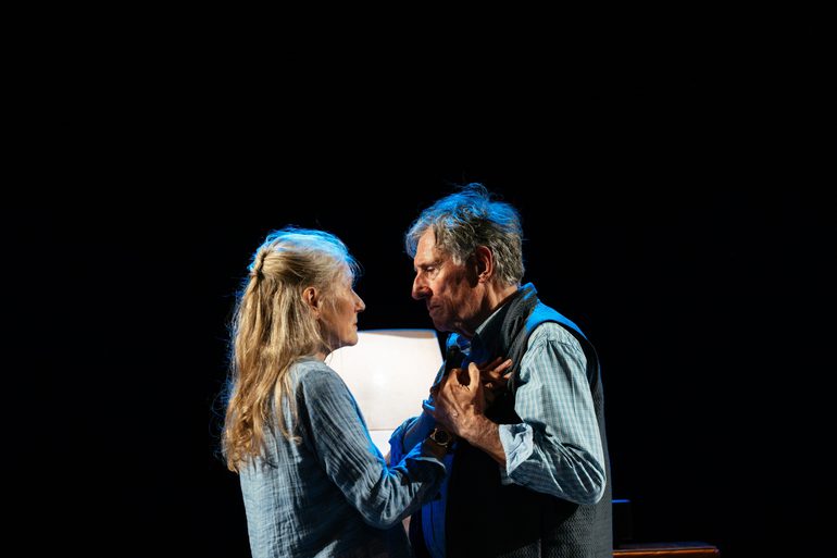 A man and a woman slow dance, looking into each others eyes with tenderness. The woman wears a blue shirt, the man wears a blue checked shirt and a gilet.