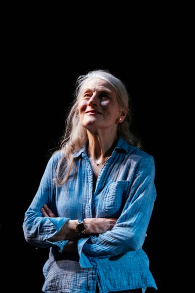 An older woman wearing a blue shirt is stood with her arms crossed. She looks out in front of her with a contented smile.