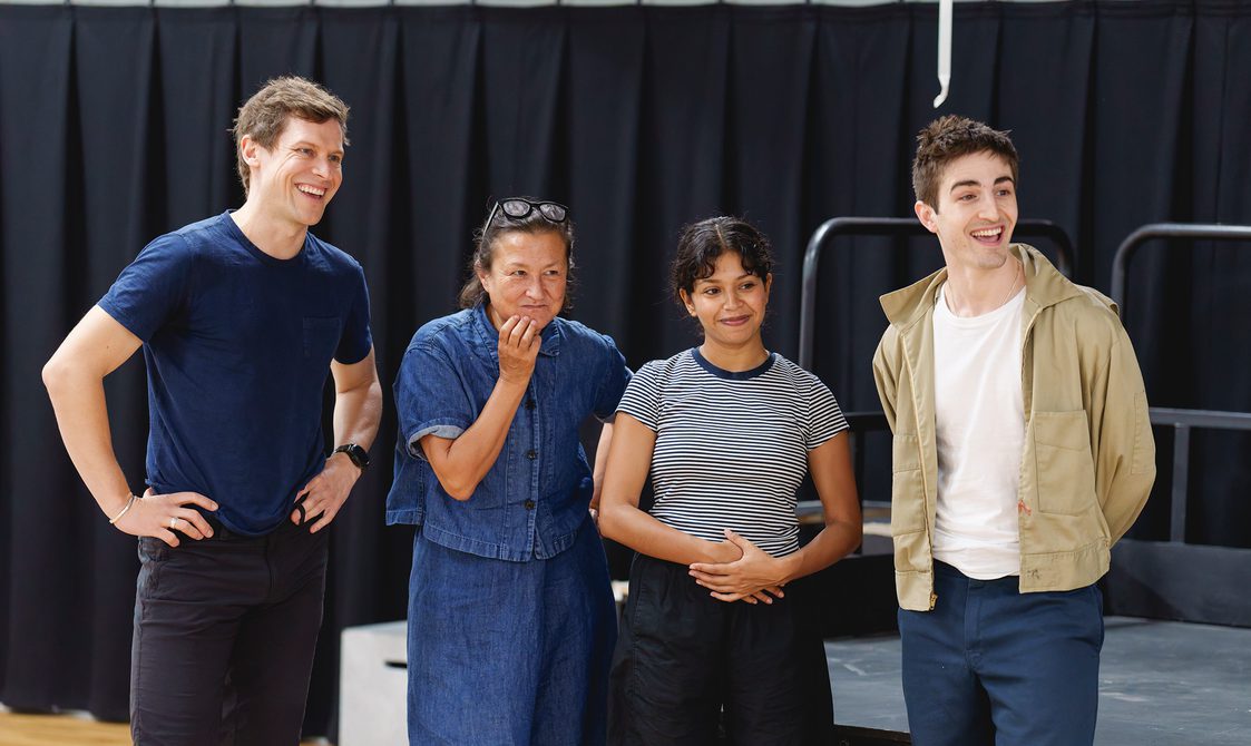 Four members of the company stand in a line. They are all looking to their left and smiling. The man on the left wears a blue t-shirt and black shorts, with his hands on his hips. The woman next to him is wearing a denim top and trousers, glasses on her head and has her hand on her chin. The woman next to her is wearing a stripy t-shirt and black trousers, her hands together in front of her. The man on the right has a white t-shirt, beige jacket and blue trousers with his hands behind his back.