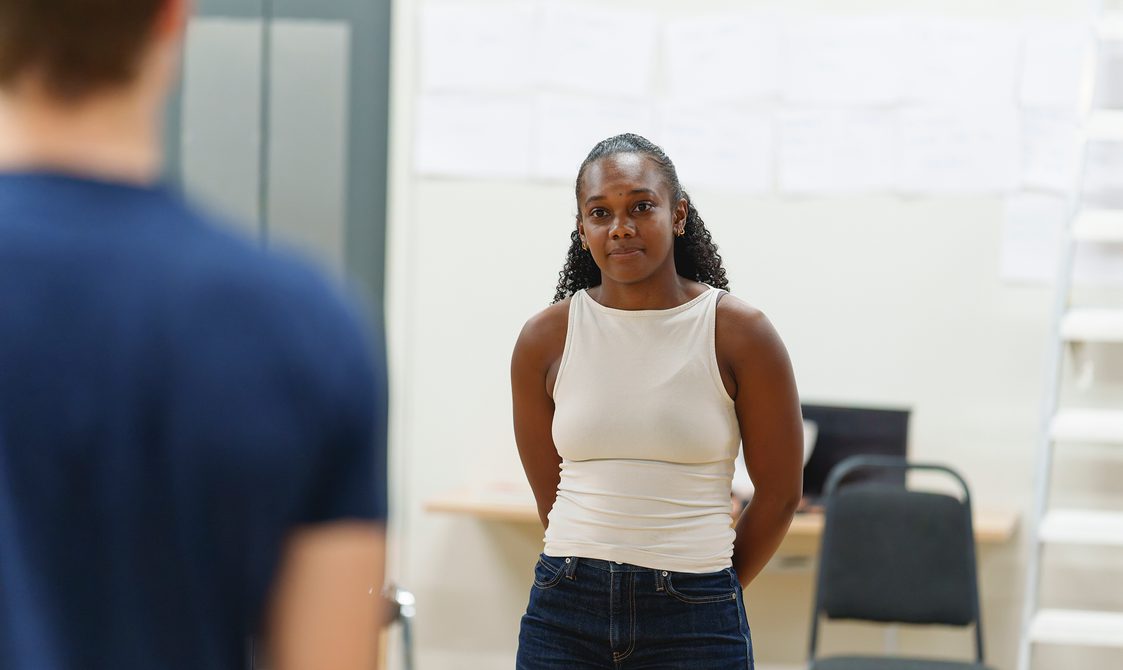 A woman with dark curly hair and a white top and blue jeans stands facing a man with his back to the camera. She has a slight smile. She holds her hands behind her back.