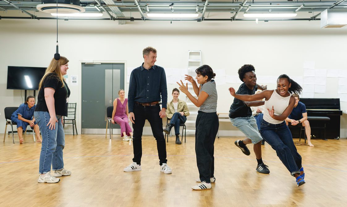 A company are mid-rehearsal. Two actors on the right are chasing each other, running. The two actors in the middle are stood still, the man is looking at the pair chasing each other and the woman is gesturing with her hands. The woman on the right stands laughing looking at the group. Other members of the company sit on chairs behind them, watching.