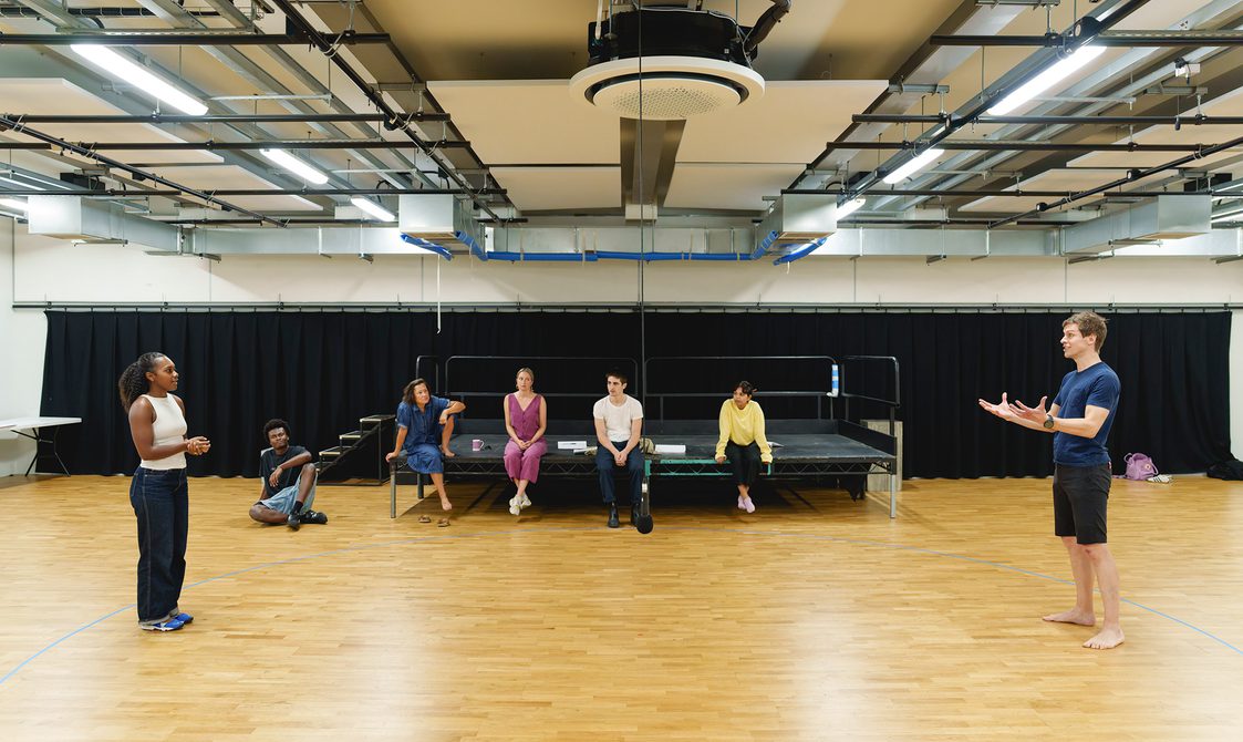A wide shot of a rehearsal studio. A man and a woman stand far apart at either end of the room. The woman has her curly hair tied up and is wearing a white top and blue jeans. She has her hands clasped together in front of her. The man is wearing a blue t-shirt and black shorts. He has his hands in front of him, palms facing up like he's holding something. Other members of the company sit on the stage behind them, watching.