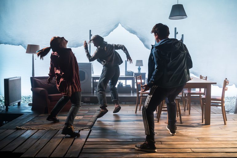 One man and two women dance erratically. Heads thrown back, arms out to the side, two of them jumping off the floor. They are in a cosy home, with wood flooring, a radiator, table and chairs, an armchair, a rug, a washing machine and lamps. A bright white background illuminates them softly.