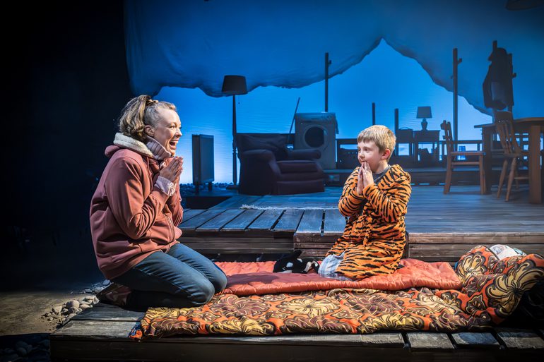 A woman and a boy kneel at the front of the stage on sleeping bags. The young boy is dressed in a tiger dressing gown, the woman has jeans and a zip-through hoodie on. They both have their hands together in front of their chest. The woman has a wide smile on her face. A house set is behind them in a blue wash.