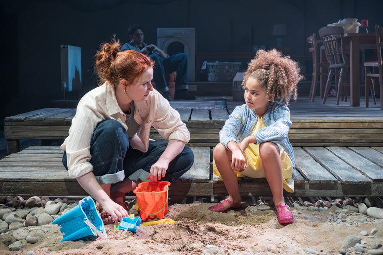 A woman and a young girl sit on a raised wooden stage, with their feet in the sand. The woman has buckets and spades in front of her. Her sleeves and trousers are rolled up, her hair tied back. She looks at the young girl with a worried expression. The young girl, in a yellow dress looks back at the woman. Behind, in a blue wash, a man sits in an armchair.