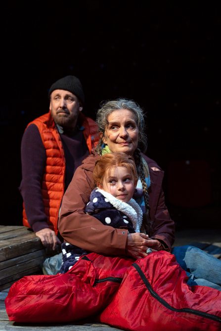A man, a woman and a young girl sit huddled together on the floor. They are dressed in hats, scarves and coats. They all look off to the side. They are in a spotlight.
