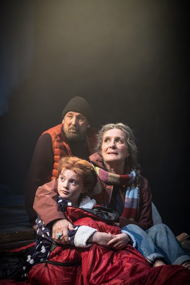 A man, a woman and a young girl sit huddled together on the floor. They are dressed in hats, scarves and coats. They all look off to the side. They are in a spotlight.