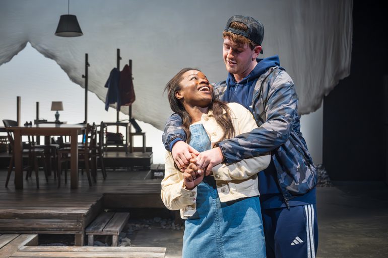 A man hugs a woman from behind. The woman looks up at him with a wide smile as he smiles down at her. A dining room scene is behind them, unlit.