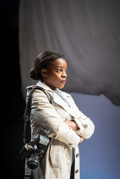A woman, in a trench coat, stands with her arms crossed. Her hair is in a bun and has a serious look on her face. She has a camera hanging over her shoulder.