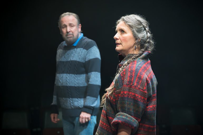 A woman stands looking off the side, her hair in a plait and the sleeves of her jumper rolled up. She has a worried expression on her face. A man is looking at her, also with a worried expression on his face.