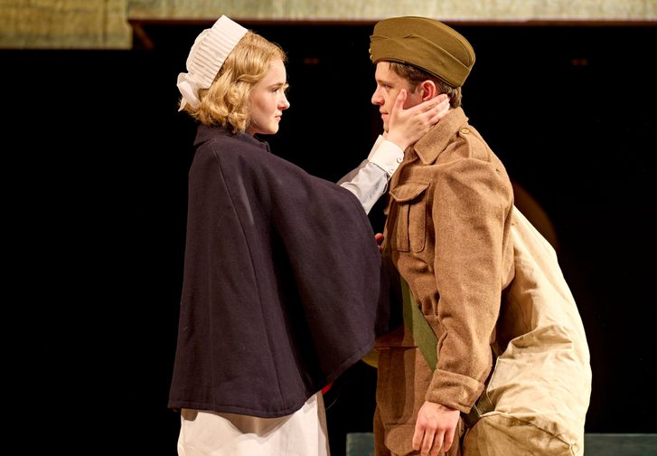 A woman wearing a nurse uniform gently holds the face of the man in front of her in an army uniform. He has a bag over his shoulder. The two look deeply at each other.