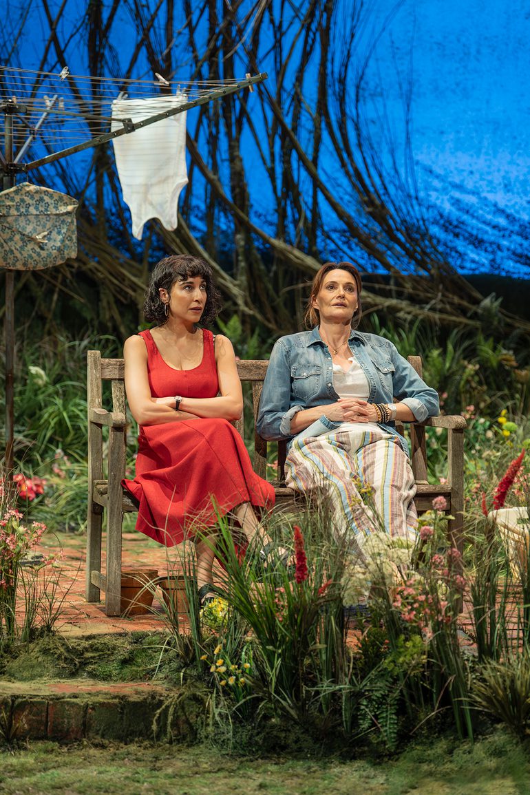 Two women sit together on a garden bench, surrounded by flowers and grass with part of a washing line in view behind them. The woman on the left wears a red summer dress and hoop earrings and has her arms crossed. The woman on the right has her medium brown hair tied up messily and wears a denim shirt and stripey linen trousers. They both look up with tired, worried expressions.