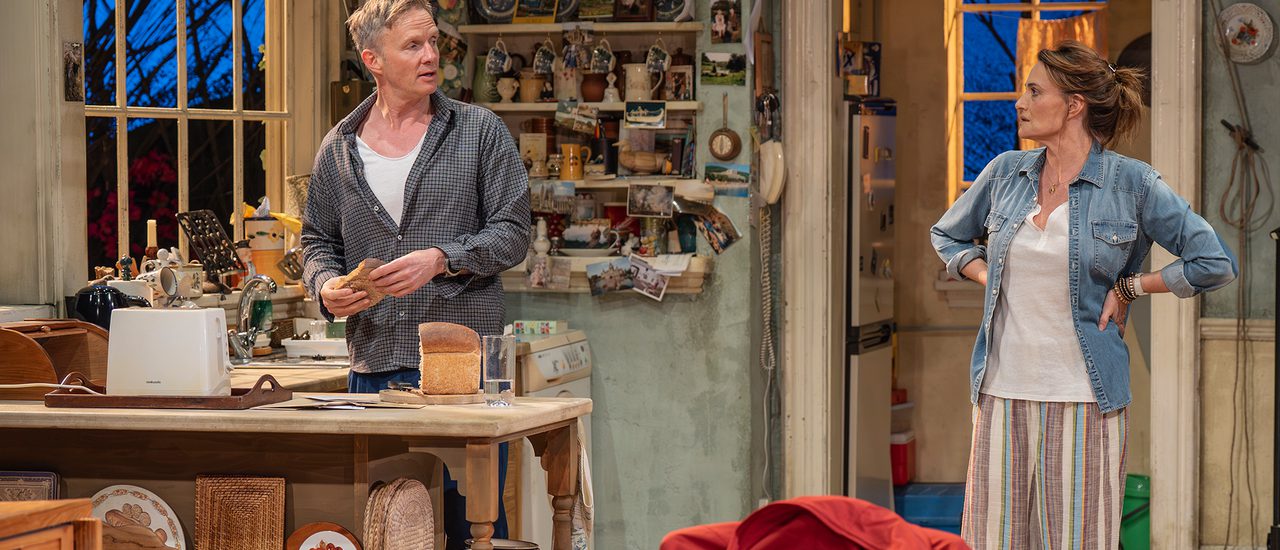A man and a woman stand talking in a cluttered farmhouse kitchen. The man is holding a slice of bread, ready to go in the toaster, and talking to the woman who stands with hands on hips looking concerned. In the foreground, a ring binder sits on the wooden dining table.