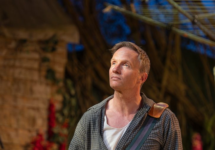 A man with short, light blonde hair stands with his hands in his pockets looking pensive. He looks up into the distance and wears a grey checked shirt, dark blue chinos and a white t-shirt. In the background is a garden full of flowers, with a shed and washing line.