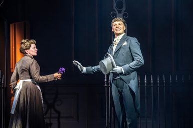 A man in coat tails and gloves smiles. He has his hat in his hand as he raises his hand up to a woman in a long brown dress and apron. She is holding a purple flower and is looking at the man. They are stood in front of railings.