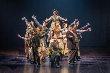 A company of actors all face outwards in a group. They all have their arms outreached, some with cups in their hands. They are all smiling widely. A man in the centre is kneeling, his hat in his hands. They wear Edwardian working-class costumes.