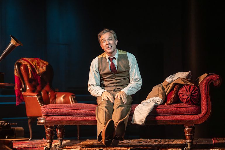 A man in a waistcoat and red tie sits on a red chaise lounge. His jacket has been thrown next to him. He stretches out his legs in front of him. A red arm chair is behind him and rugs cover the floor.