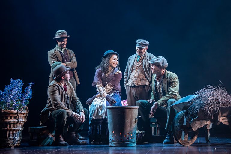 A woman with curly brown hair and a hat sits in the centre of four men. There is a metal bin, glowing orange from inside in front of them. A basket of flowers and a wheel barrow of straw are either side of the gorup. The men are looking and smiling at the women as she speaks to them. Two are sitting, the other two are standing and have their arms crossed and the other with his hands in his pockets.