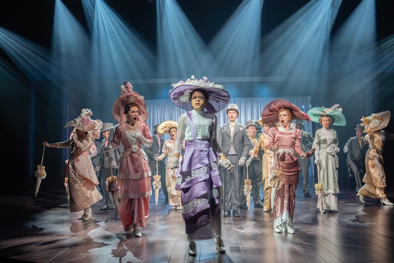 A woman in a purple dress and fascinator stands at the front of the stage. She has a defiant look, her eyebrows scrunched. The rest of the company have shocked expressions, some with their hands to their chest, others to their mouths.