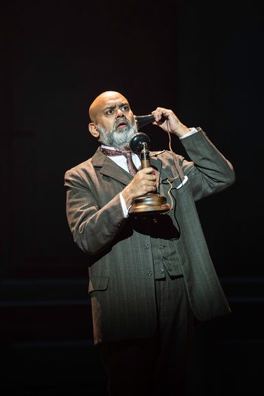 A man in a three piece, grey suit stands holding an Edwardian telephone. He is listening with a shocked expression on his face.