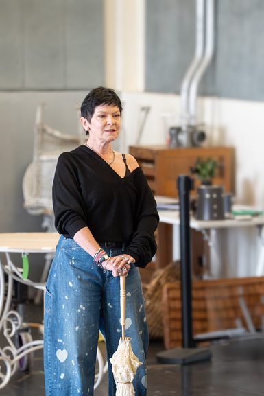 A performer wearing baggy blue jeans and a black jumper, which sits off one shoulder, stands in a studio holding a parasol, closed and resting on the floor, in both hands. She has short, dark hair and silver chunky jewellery.