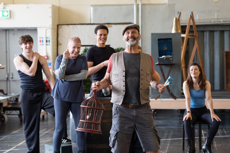 A performer with grey facial hair stands singing joyfully in a studio, holding a birdcage in one hand and a closed fan in the other. He is wearing grey cargo shorts, a grey and red t-shirt, a brown waistcoat and a flat cap. Behind him, a group of other performers sit and stand, watching on, leaning on a wooden barrel.