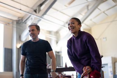 A man with grey hair and a young woman with dark, curly hair, tied in a tight bun, stand together in a studio, laughing. They are looking away from the camera.