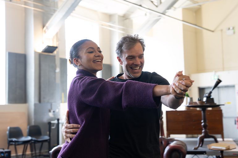 A younger woman with her dark, curly hair tied in a tight bun dances joyfully in hold with a man with grey hair in a studio. Both smile and look towards their hands which are clasped and held out straight ahead of them. They wear casual clothes.
