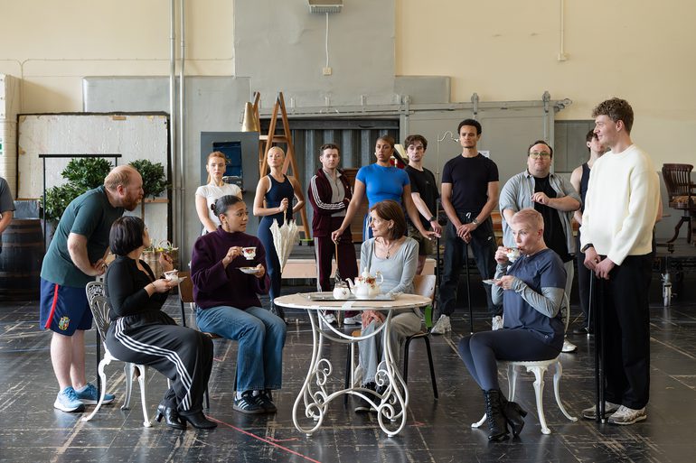 A group of performers in a rehearsal studio. In the centre, four women sit around an outdoor white café table holding traditional tea cups. The younger woman talks to the older woman in the middle, whilst the others sip their tea. Around them, a group watches on with interest, standing proudly.