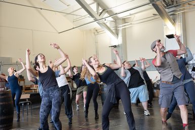 A group of performers in a rehearsal studio, midway through a dance number. They are all moving together, with their fingers crossed and held above their heads, and they look up in the same direction. There is a joyful, energetic atmosphere.