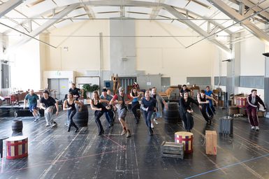 A group of performers in a rehearsal studio, midway through a dance number. They are arranged in lines, crossing one leg over the other, with their arms in a running position, singing enthusiastically. They wear casual, colourful clothes. Around them, a selection of wooden barrels and crates are littered across the floor.