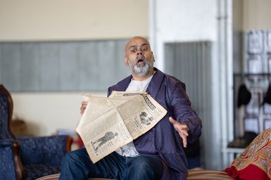 A bald man with grey facial hair sits on a chaise longue holding an old newspaper. He looks down, mouth open in amazement, with one hand outstretched in front of him. Beside him is a dark blue armchair.