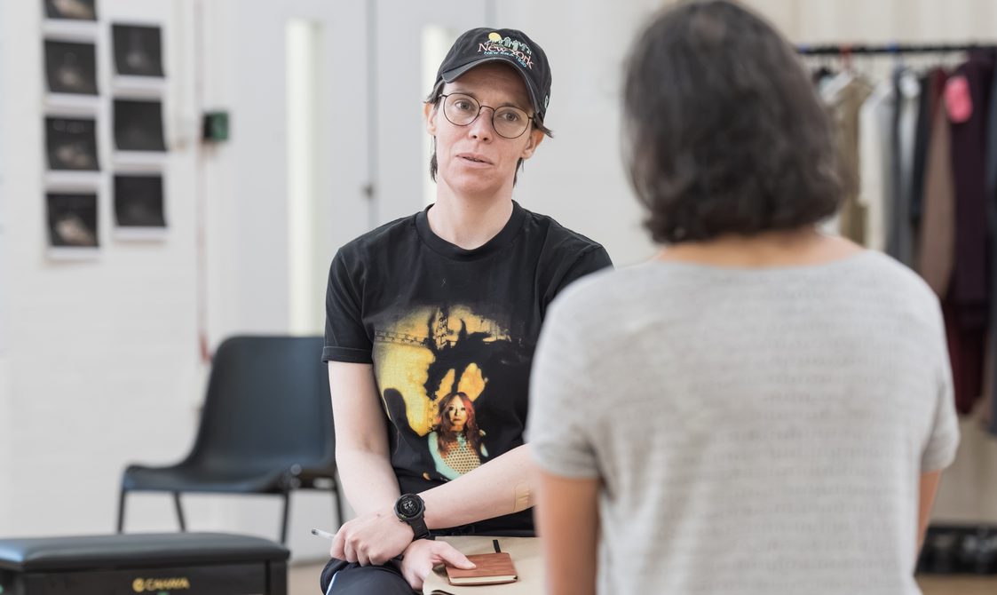 A performer with a black cap that reads 'New York' on, glasses and a black graphic t-shirt sits looking at another performer who has their back to the camera, out of focus. The perfomer has a notebook on their lap and a pen in their hand.