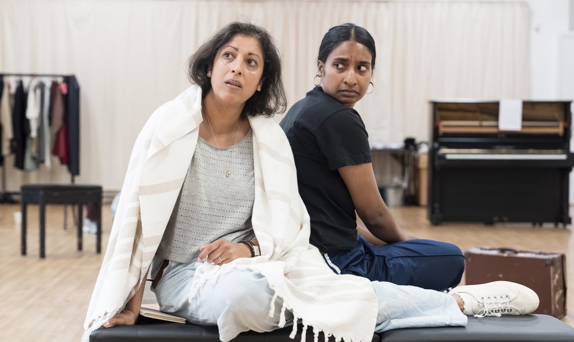 Two women sit on a bench seat. The woman on the left has her feet up on it and is wearing a blanket around her shoulders. She has short dark hair, a grey t-shirt and light wash jeans. She is looking to the side in wonder. The woman next to her is sat with her back to her but her head is turned completely to look at her with a worried expression. She is wearing a black t-shirt and navy trousers. They are in a rehearsal studio.