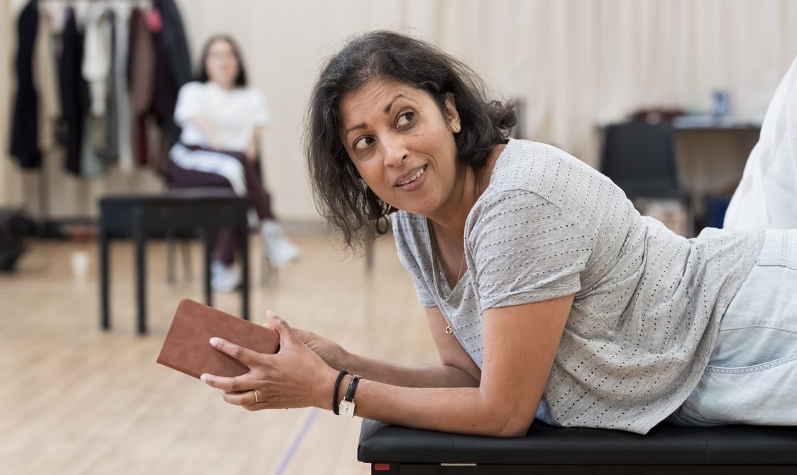 A woman with short dark hair is lying down on a bench. She has a book in her hands and is looking behind her. She is smiling. Behind her is a rehearsal studio, out of focus. Another member of the company is sat on a chair.