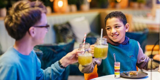 Two boys clink their glasses together at a table; they are surrounded by sweet treats on the table.